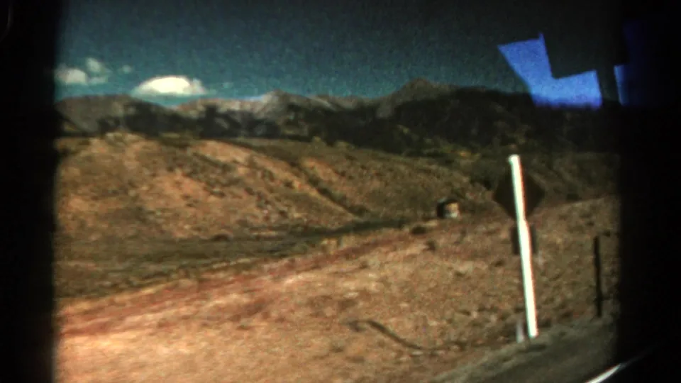 1967 Colorado 8mm frame with a white roadside post, dry open hillside, distant mountains, and blue window reflection.