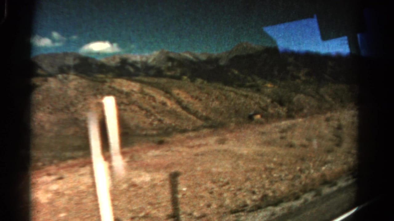 1967 Colorado home-movie view through a car window, showing rocky hills, distant mountains, scrub land, and a roadside pole.
