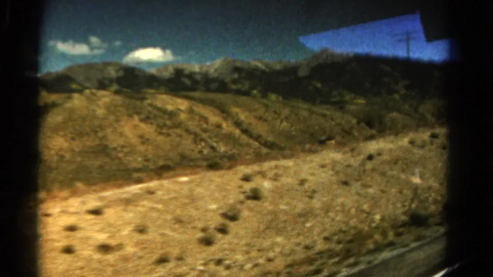 From inside a moving car, the 1967 Colorado footage shows a pale road edge, rocky scrub terrain, and distant rugged mountains.