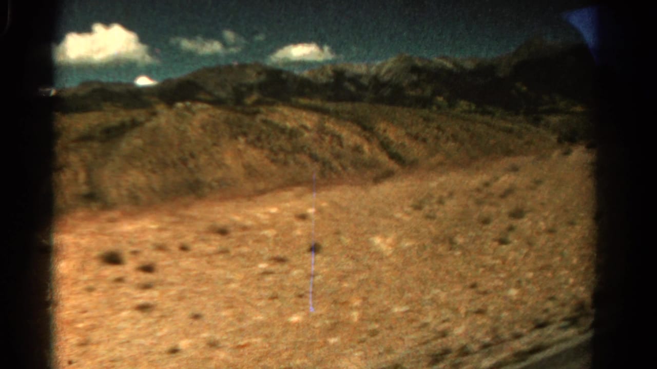 1967 Colorado 8mm frame looking across dry hills, sparse scrub, distant mountains, and a clouded blue sky from a moving roadside view.