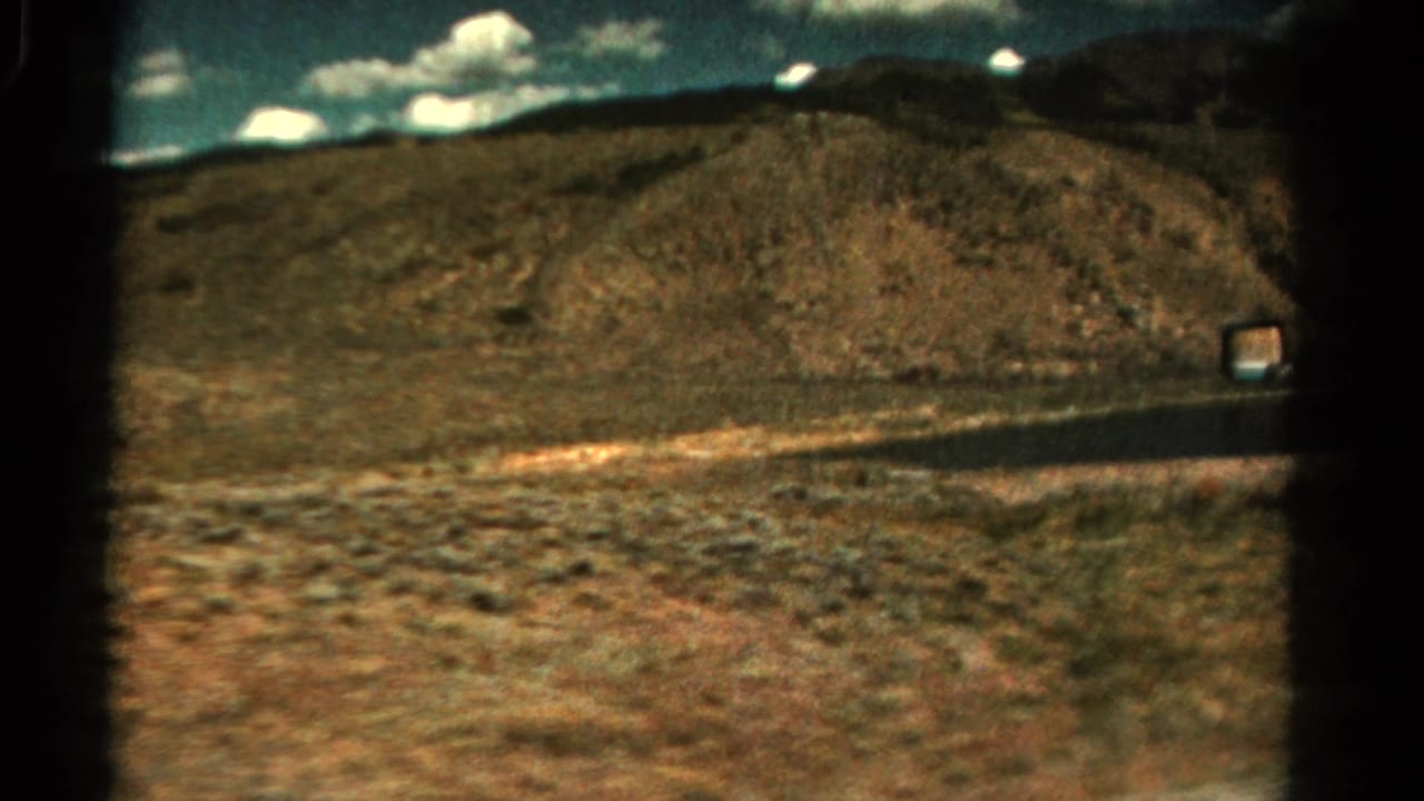 Paved road crossing dry terrain below low hills and a clouded sky.