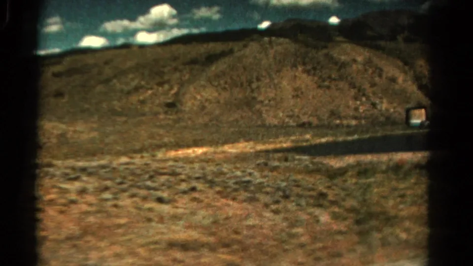 1967 Colorado roadside view with a paved road cutting across arid ground, low hills, distant mountains, and scattered clouds.