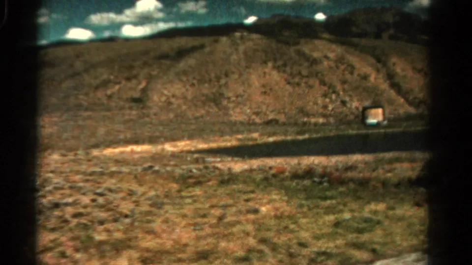1967 Colorado 8mm frame showing a small roadside structure near a paved road, with dry hills, scrub, and distant mountains behind it.