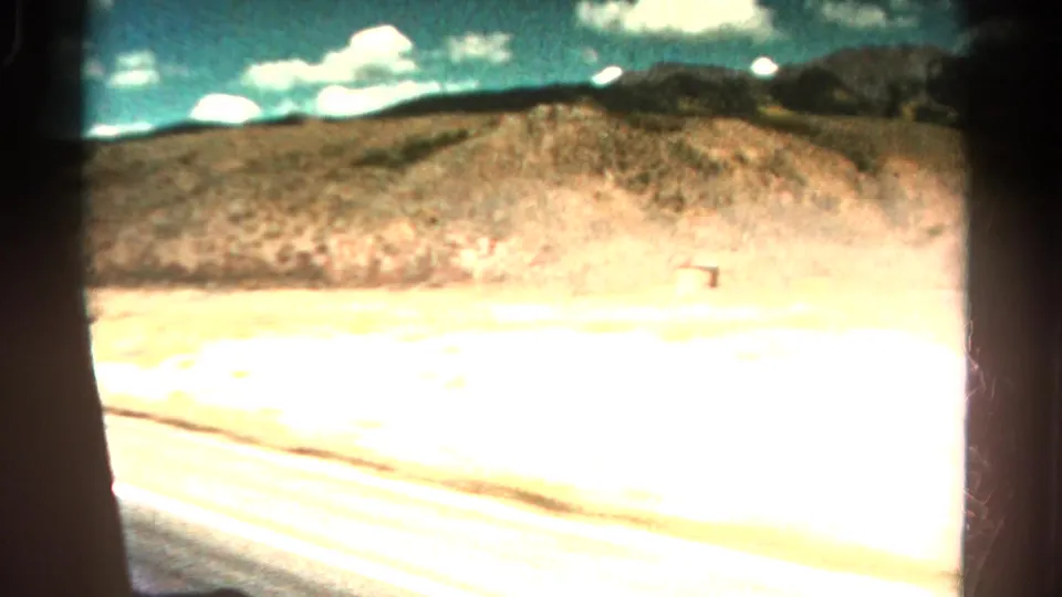 1967 Colorado travel frame with bright exposure across the road edge, dry hillside, scattered scrub, and clouds over distant mountains.