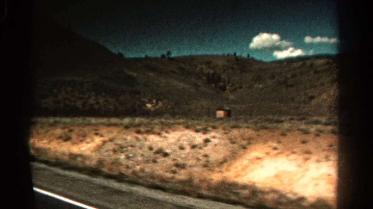 1967 Colorado 8mm frame from a moving vehicle showing a road edge, arid hillside, sparse vegetation, and white clouds.