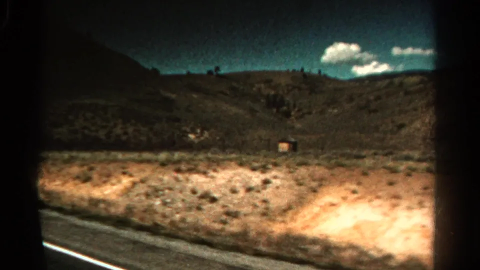 1967 Colorado 8mm frame from a moving vehicle showing a road edge, arid hillside, sparse vegetation, and white clouds.