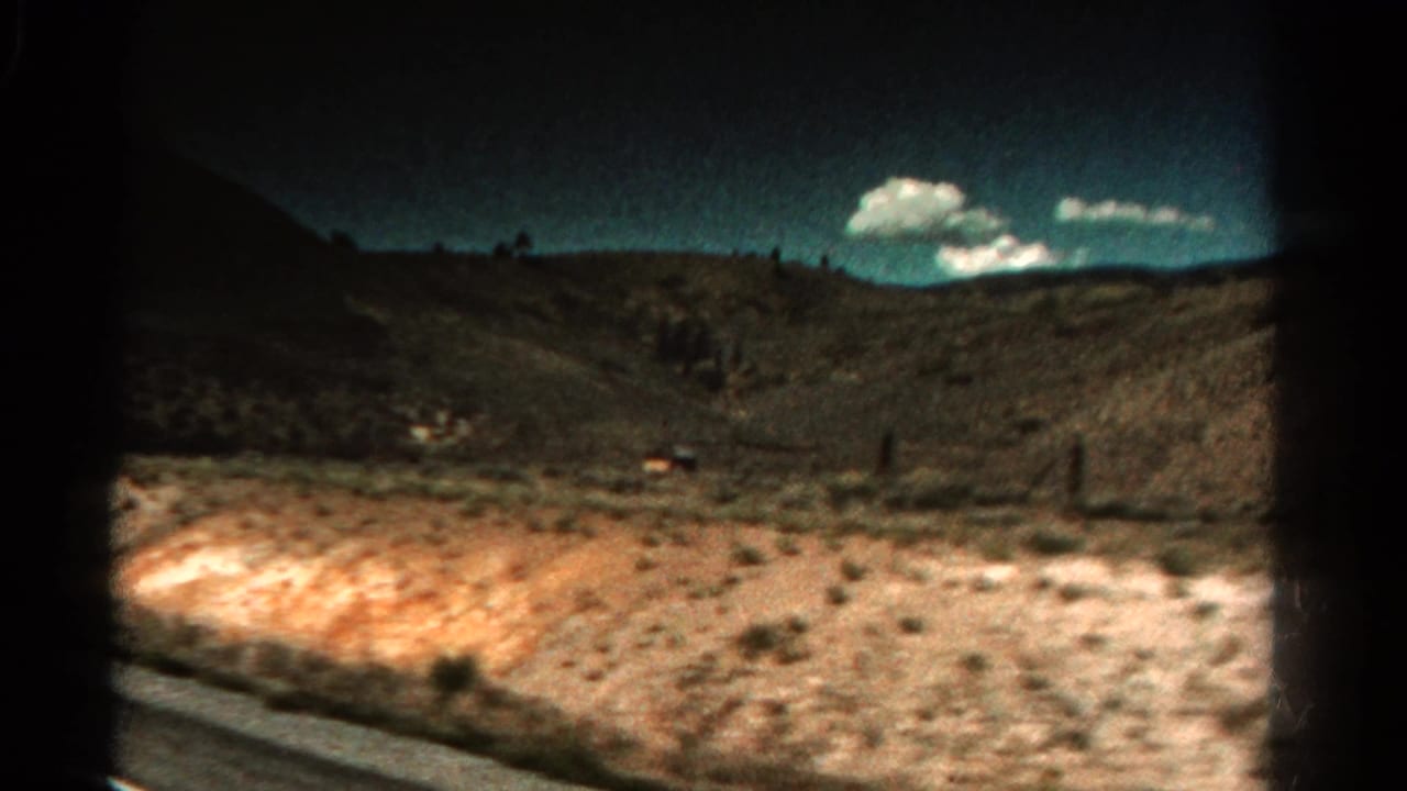 Dry roadside slope with scattered brush, distant hills, fence posts, and small white clouds.