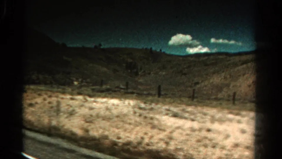 1967 Colorado road-travel frame with motion blur across a dry shoulder, fence line, low hills, and bright clouds.