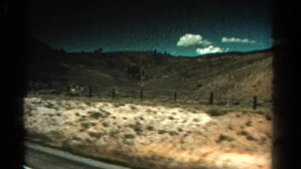 Final 8mm frame of a Colorado roadside landscape with dry brush, fence posts, low hills, and clouds from a moving vehicle.