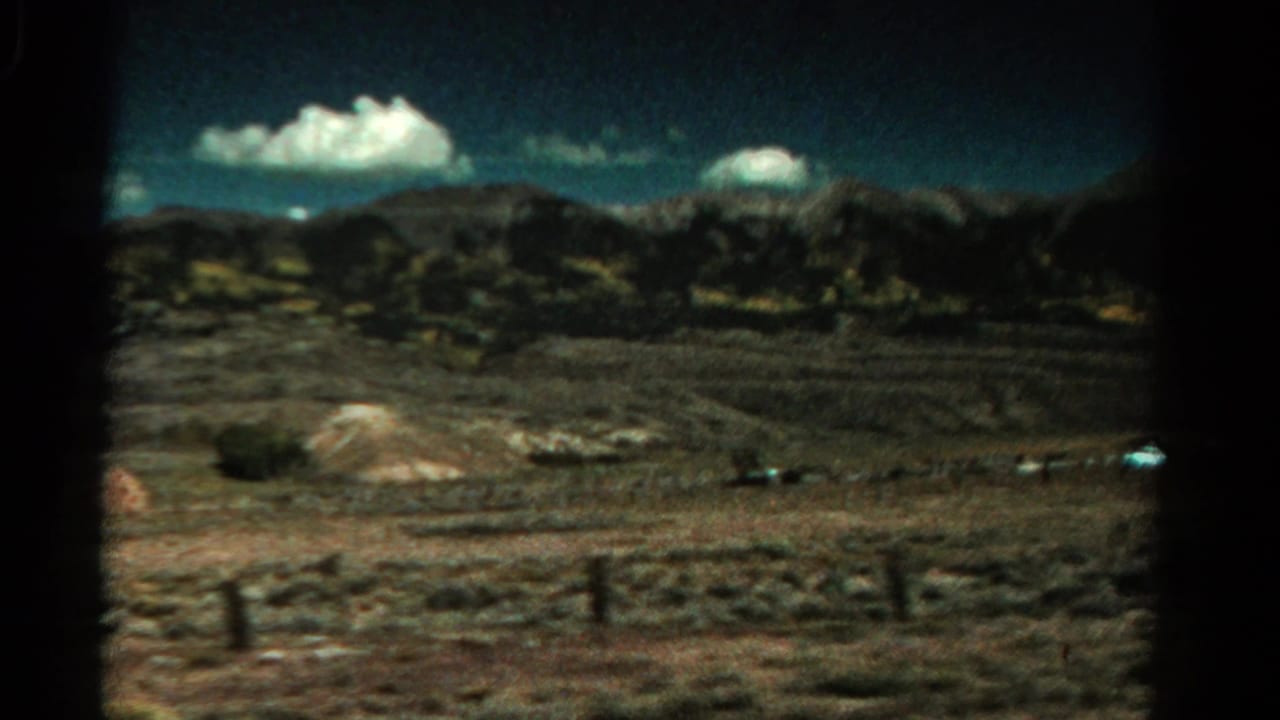 Dry Colorado countryside with fence posts, distant mountains, blue sky, and white clouds seen from a moving vehicle.