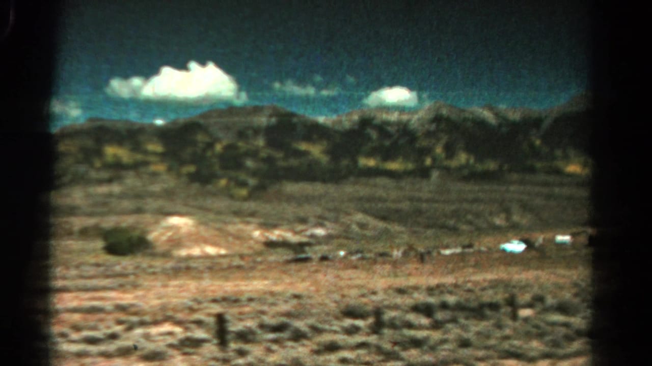 Rural Colorado landscape in 1967 home-movie footage, showing dry open ground, a fence line, distant mountains, and puffy white clouds.