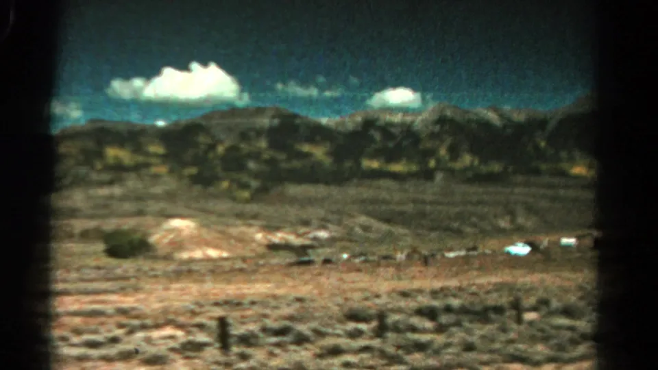 Rural Colorado landscape in 1967 home-movie footage, showing dry open ground, a fence line, distant mountains, and puffy white clouds.