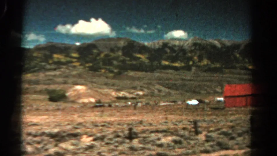 1967 Colorado countryside view with a red barnlike structure at the right edge, dry terrain, fence posts, mountains, and white clouds.