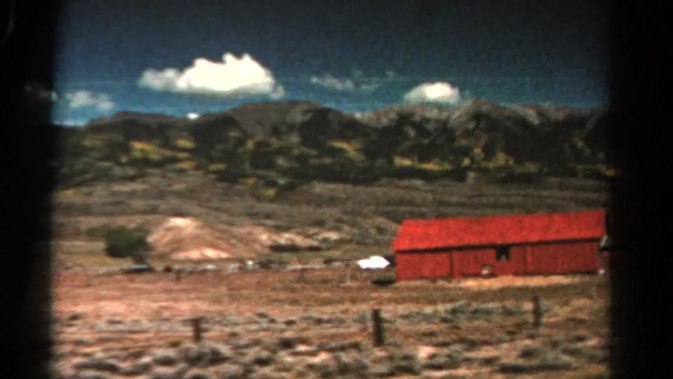 1967 8mm frame of a red barnlike building in rural Colorado, set against dry open land, fence posts, mountains, and bright clouds.