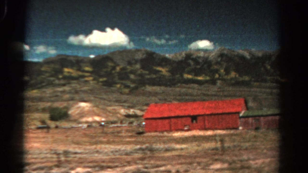 Red barn and outbuilding beside dry open ground with mountains and clouds behind.