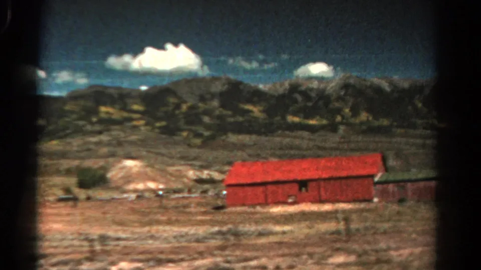 1967 Colorado 8mm frame showing red farm buildings, dry roadside terrain, distant mountains, and bright white clouds.