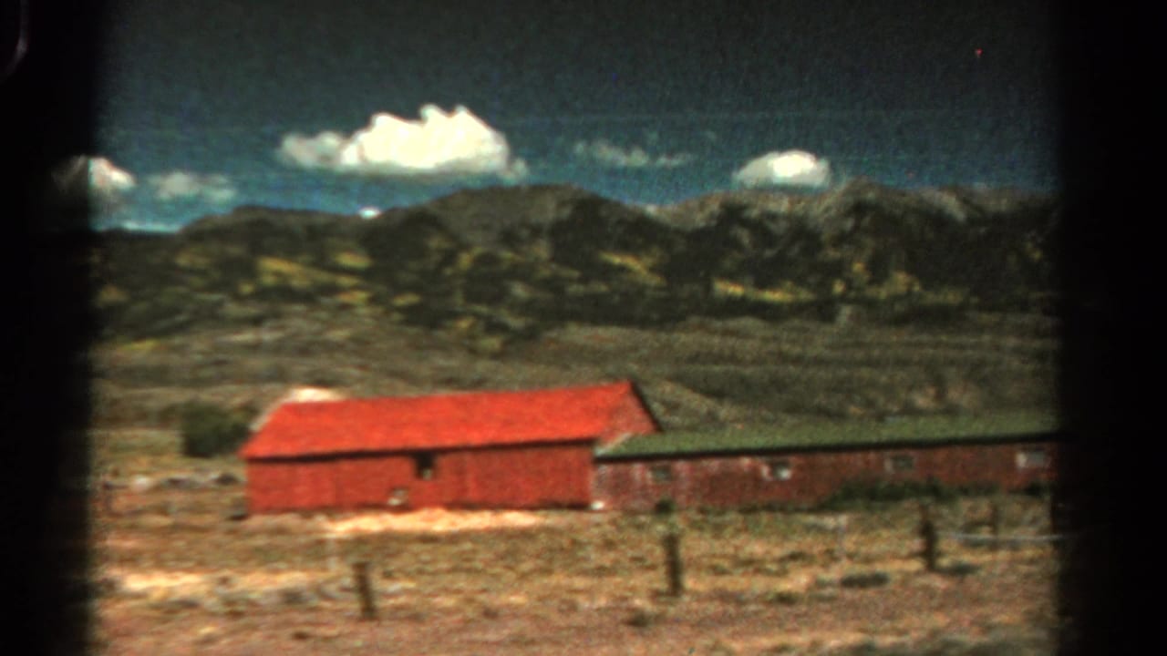 Red barn and long low building in open terrain with mountains and clouds beyond.