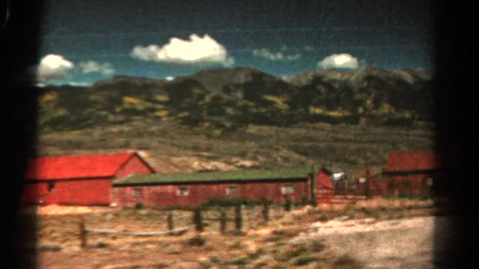 1967 Colorado 8mm frame with red roadside buildings, fence posts, dry terrain, and mountains under a clouded sky.