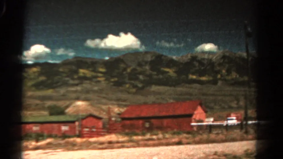 1967 Colorado home-movie frame showing red barns near a road, open dry ground, mountain ridges, and bright clouds.