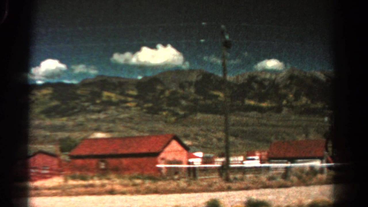 1967 Colorado home-movie frame showing red-roofed roadside buildings, a white fence, utility pole, open land, and distant mountains.