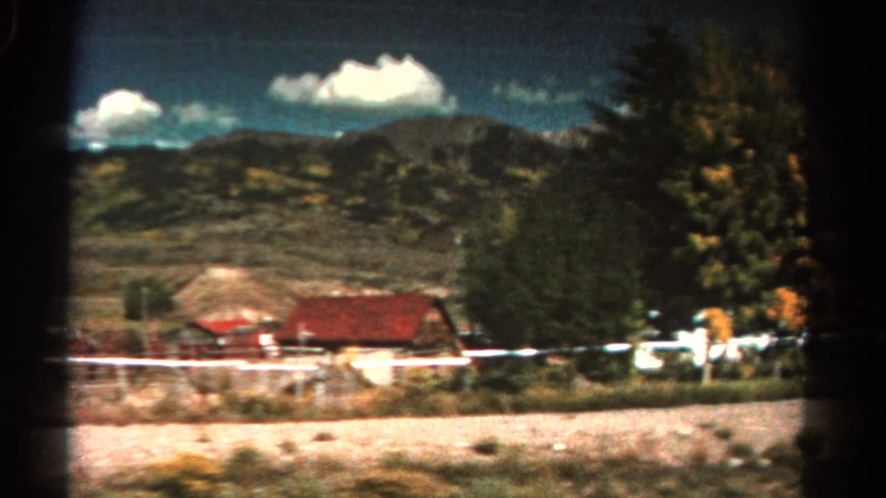 Roadside houses, white fencing, trees, and mountains seen from a moving vehicle.