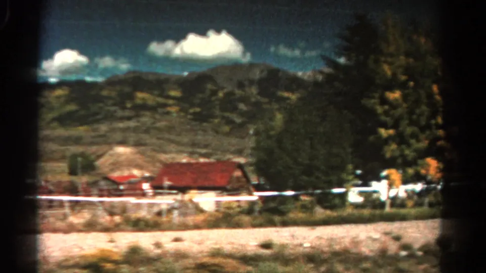 1967 Colorado roadside view with low houses, white fencing, trees, open ground, and mountain ridges under scattered clouds.