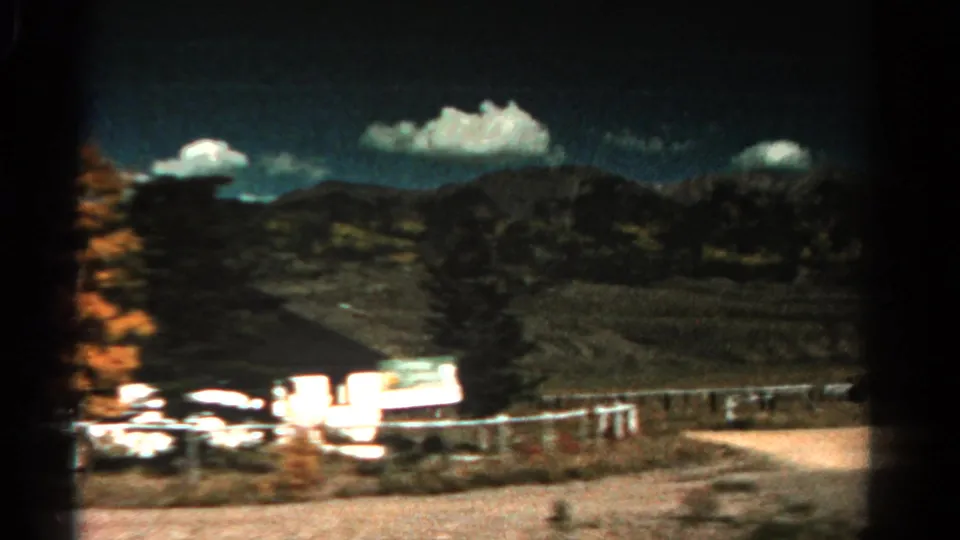 1967 Colorado driving view showing a small white roadside structure, fencing, open fields, dark mountain slopes, and bright clouds.
