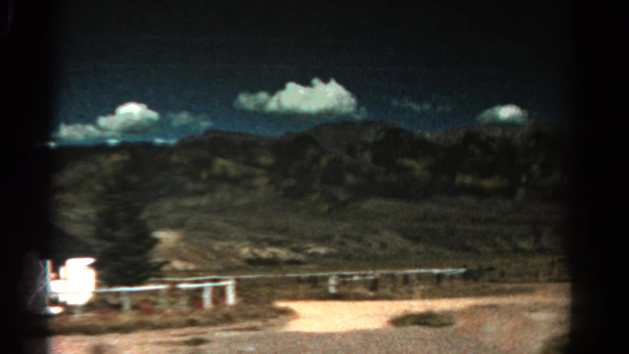 Dry roadside landscape with low mountains, blue sky, white clouds, and a pale fence near the road.