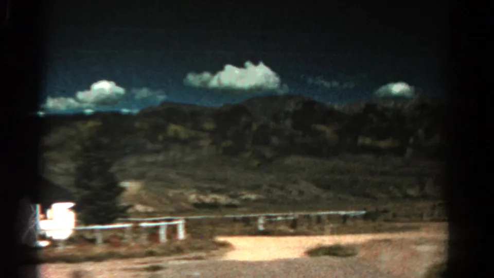 1967 Colorado 8mm frame showing an arid roadside view with distant ridges, scattered clouds, fencing, and dark film gate edges.