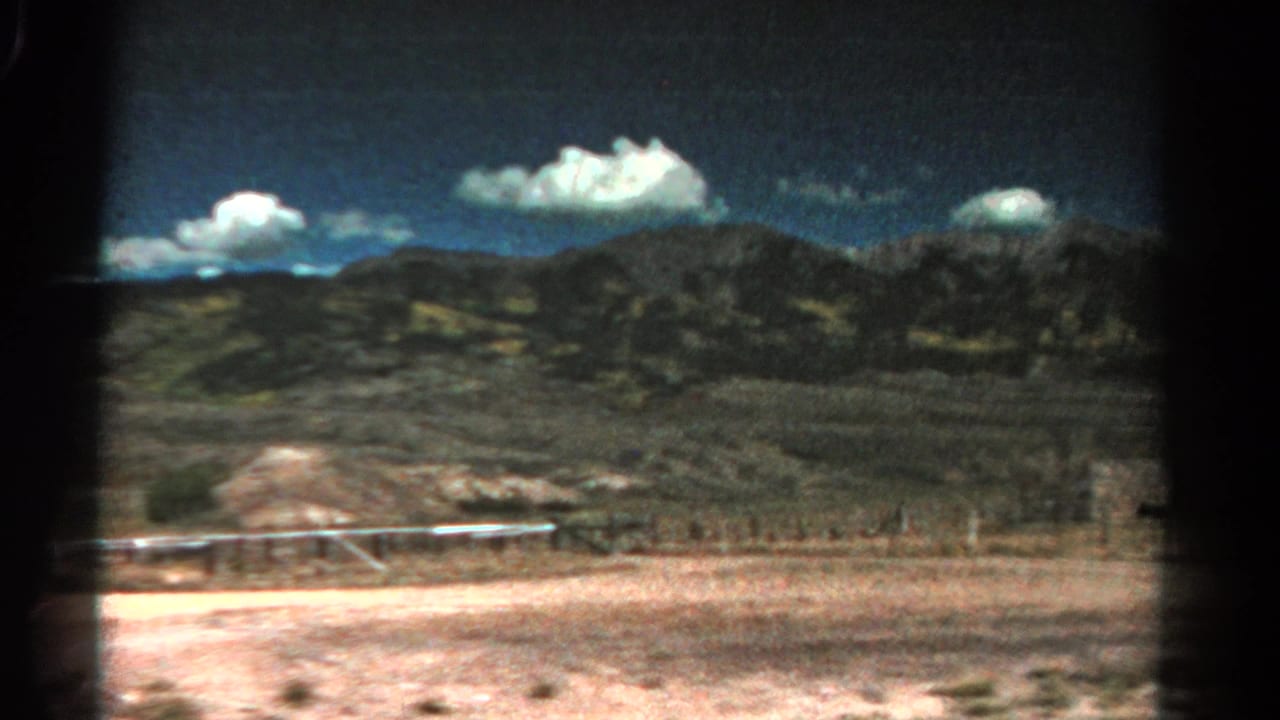 1967 Colorado road-view frame with dry foreground, a low fence line, distant mountain slopes, and cumulus clouds on 8mm film.