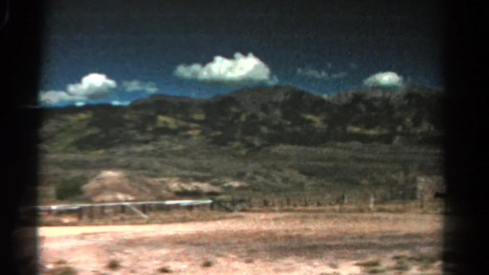 1967 Colorado road-view frame with dry foreground, a low fence line, distant mountain slopes, and cumulus clouds on 8mm film.