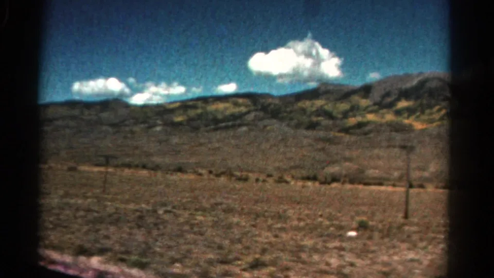 1967 Colorado 8mm frame of a broad arid plain, distant ridges, roadside utility pole, and bright clouds under a blue sky.