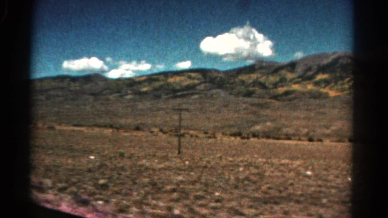 1967 Colorado home-movie frame of arid countryside and low hills, with a utility pole in the midground and clouds overhead.