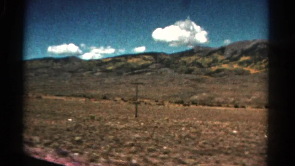 1967 Colorado home-movie frame of arid countryside and low hills, with a utility pole in the midground and clouds overhead.