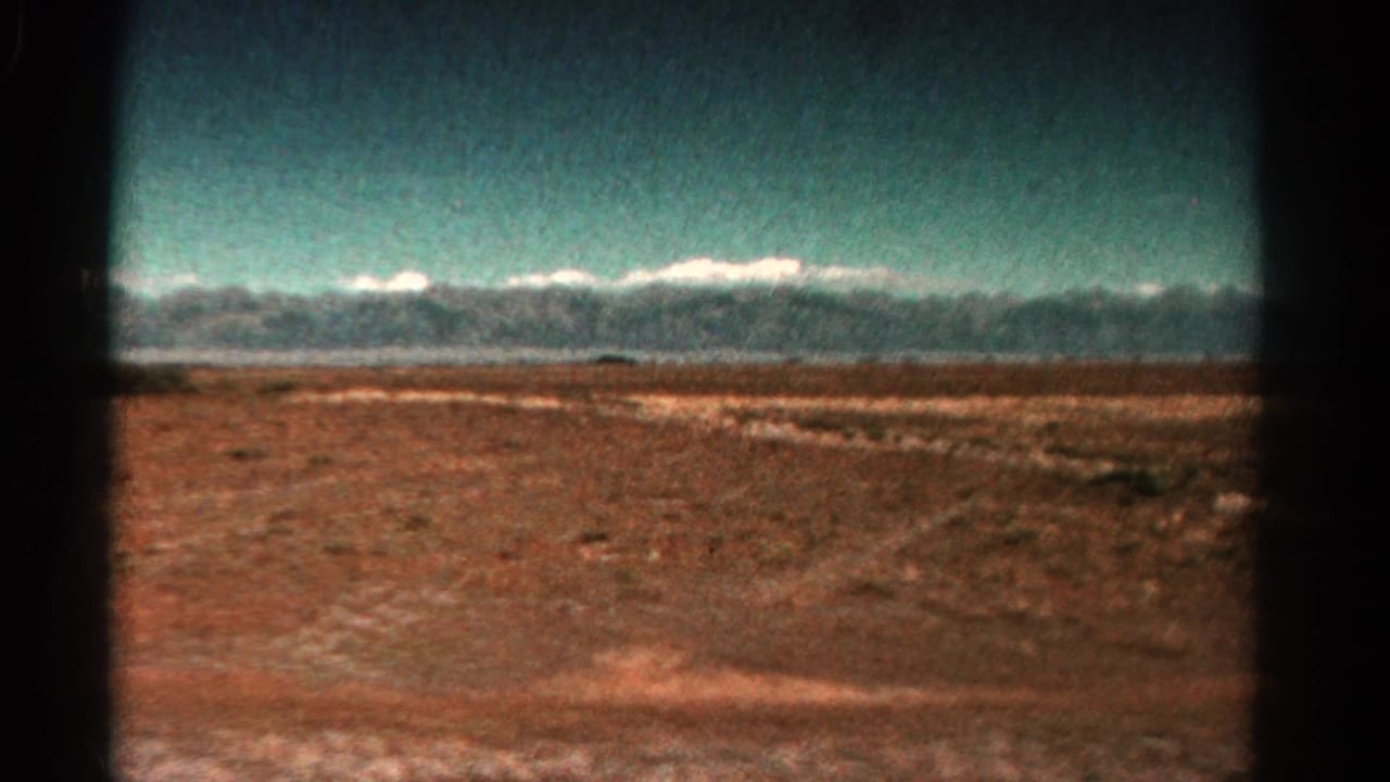 1967 Colorado 8mm frame showing an arid plain, scattered vegetation, and a distant snow-capped mountain range.