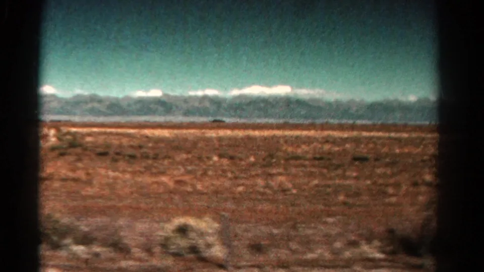 1967 Colorado home-movie frame showing reddish arid terrain, sparse vegetation, and a distant snow-capped range.