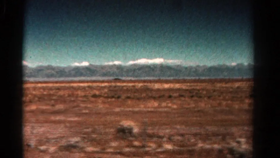 1967 Colorado 8mm frame showing a long mountain horizon beyond arid roadside land, softened by film grain and vehicle motion.