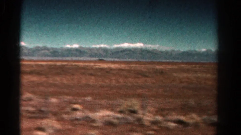 1967 Colorado home-movie frame with reddish scrubland in front of a distant snow-capped range, bordered by dark 8mm gate edges.