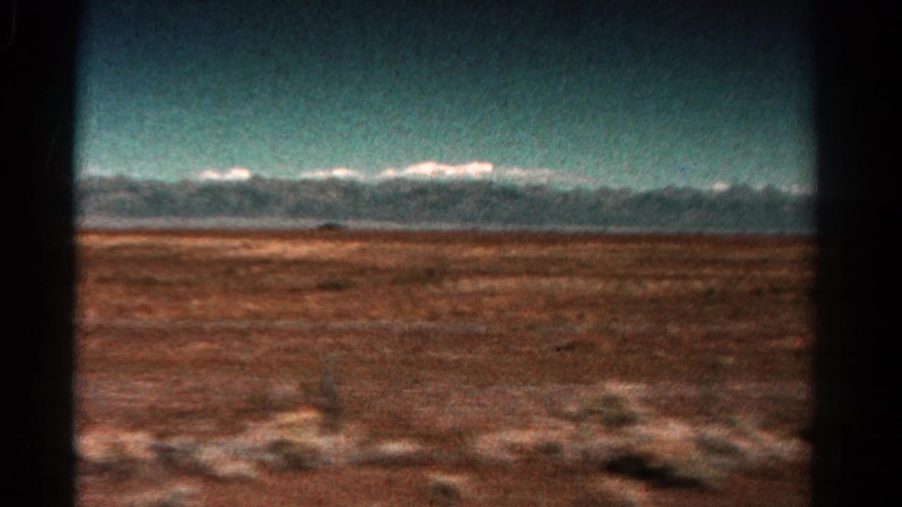 Distant snow-capped mountains beyond reddish open terrain, framed by a dark vehicle window.