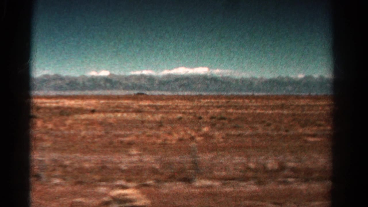 1967 Colorado road-view frame with reddish ground, distant snow-capped peaks, teal sky, and dark vehicle interior edges.