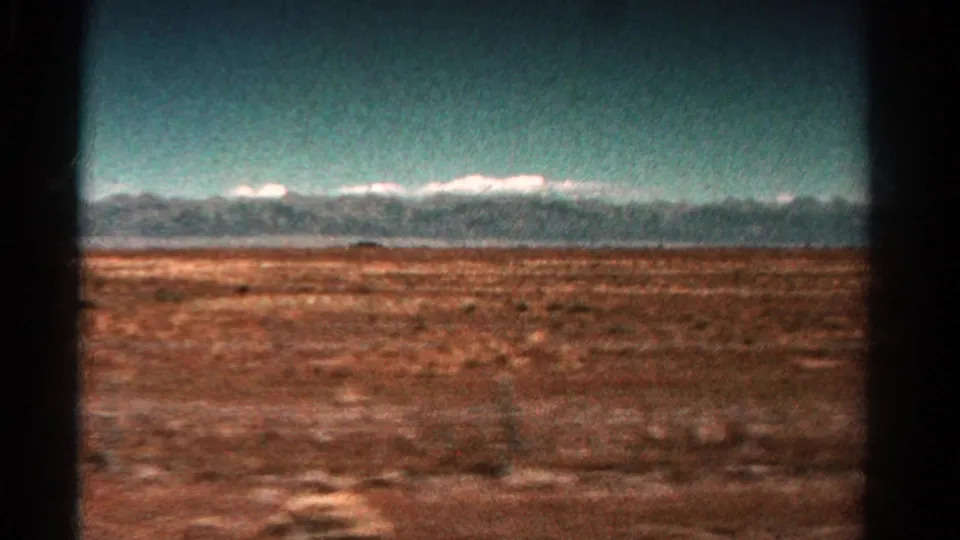 1967 Colorado road-view frame with reddish ground, distant snow-capped peaks, teal sky, and dark vehicle interior edges.