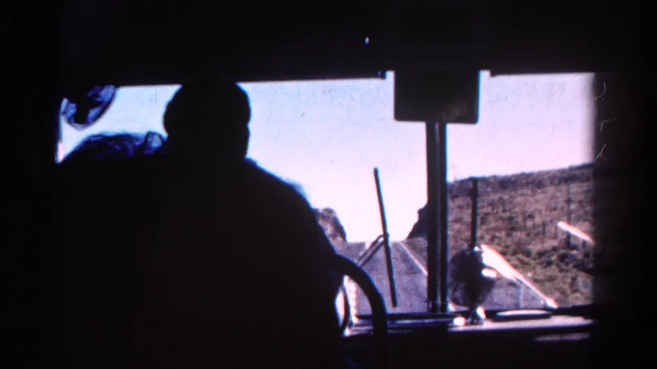Interior vehicle view in 1967 Colorado, with a silhouetted passenger, windshield hardware, road ahead, and open arid landscape outside.