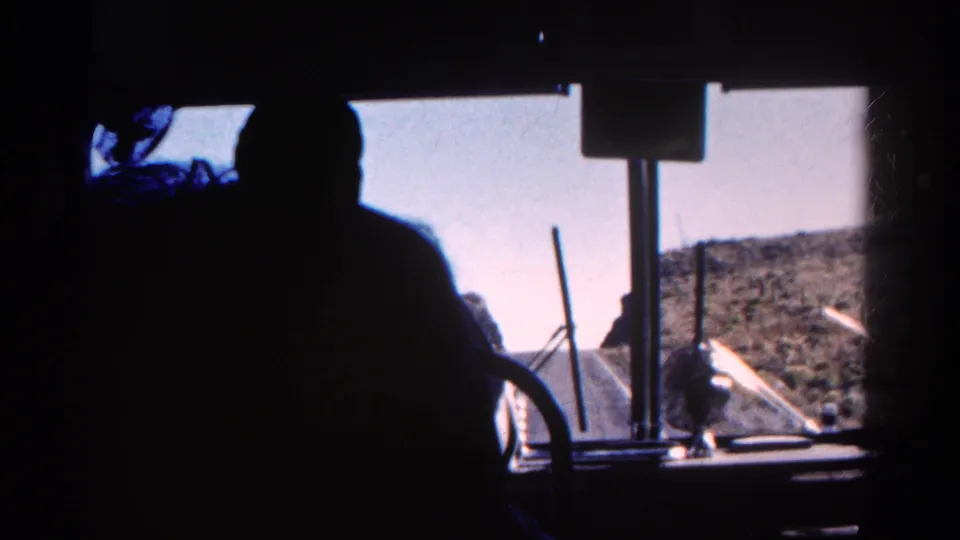 Inside-cab view from 1967 Colorado footage, showing a dark human silhouette, windshield post, wipers, bright sky, and rugged hillside.