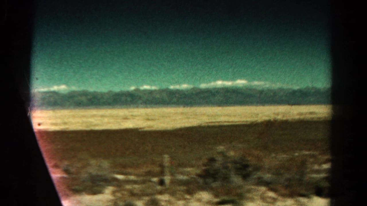 Train-window view of flat Colorado lowland with distant mountains and teal sky.