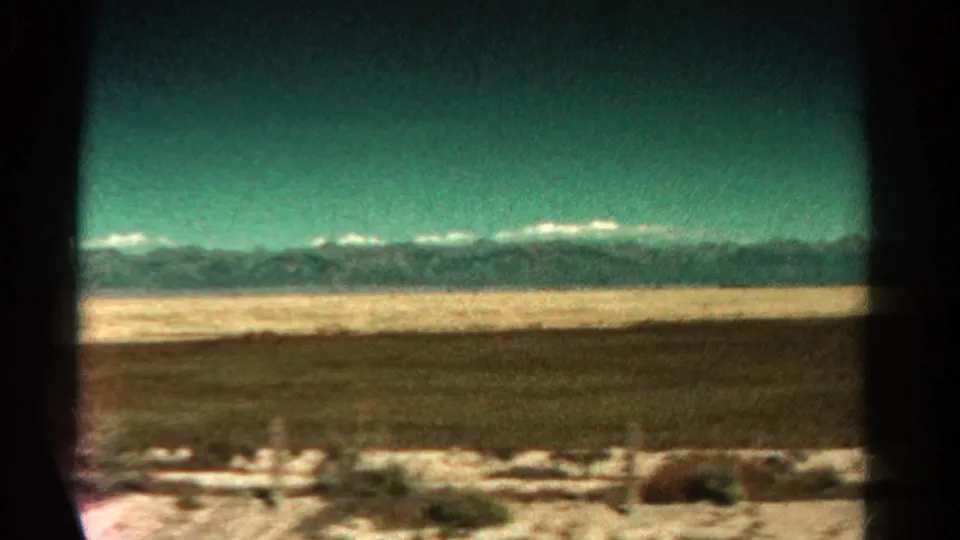 1967 home-movie frame from a moving train, with blurred foreground brush and distant Colorado mountains beyond flat terrain.