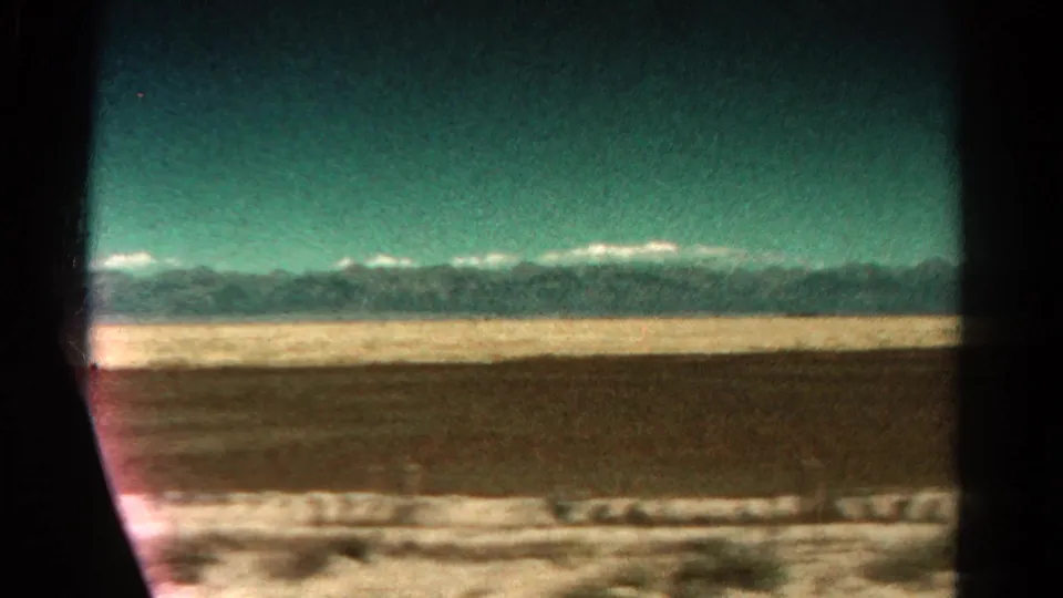 Passenger-train window view in Colorado, with dark frame edges, open lowland, sparse vegetation, and mountains on the horizon.