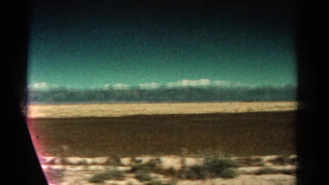 1967 Colorado road-view frame with snow-capped mountains beyond an open plain, seen through the edge of a moving vehicle window.