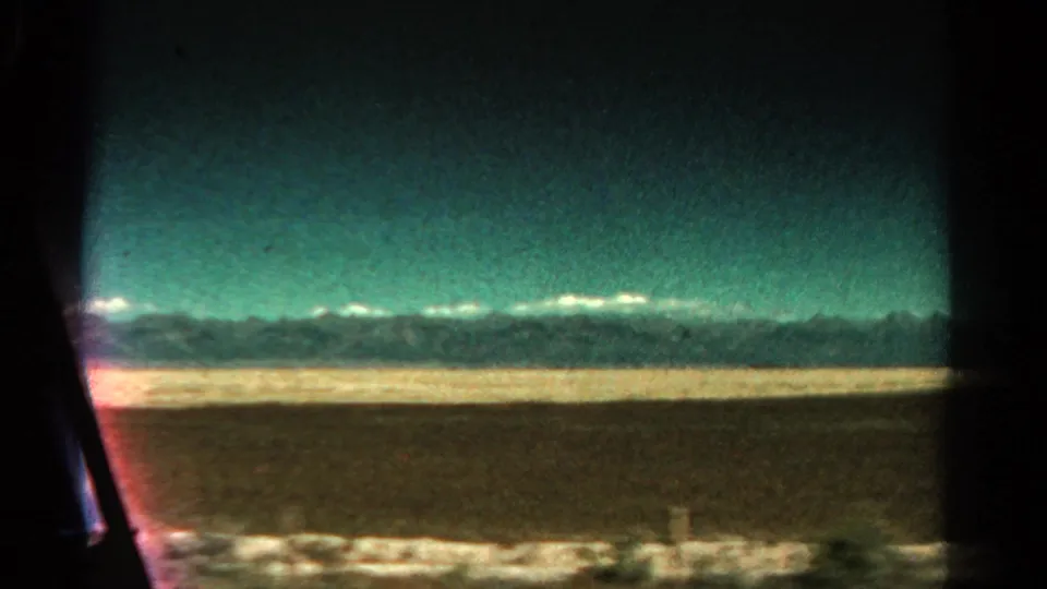 Distant Colorado mountains and a flat brown plain pass by from inside a moving vehicle in this 1967 8mm frame.