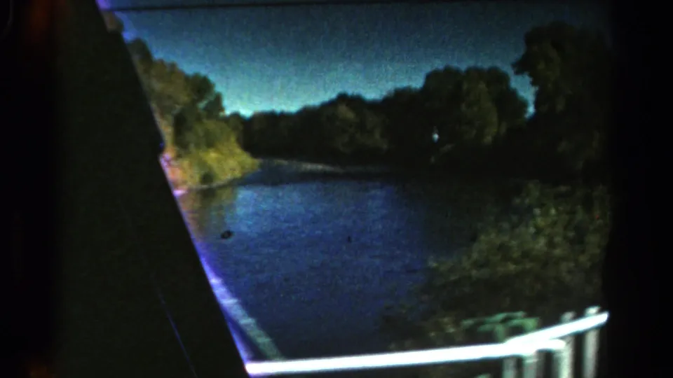 Colorado 1967 home-movie frame showing water, a tree-lined shore, darkening sky, and a bright rail or vehicle edge in motion.