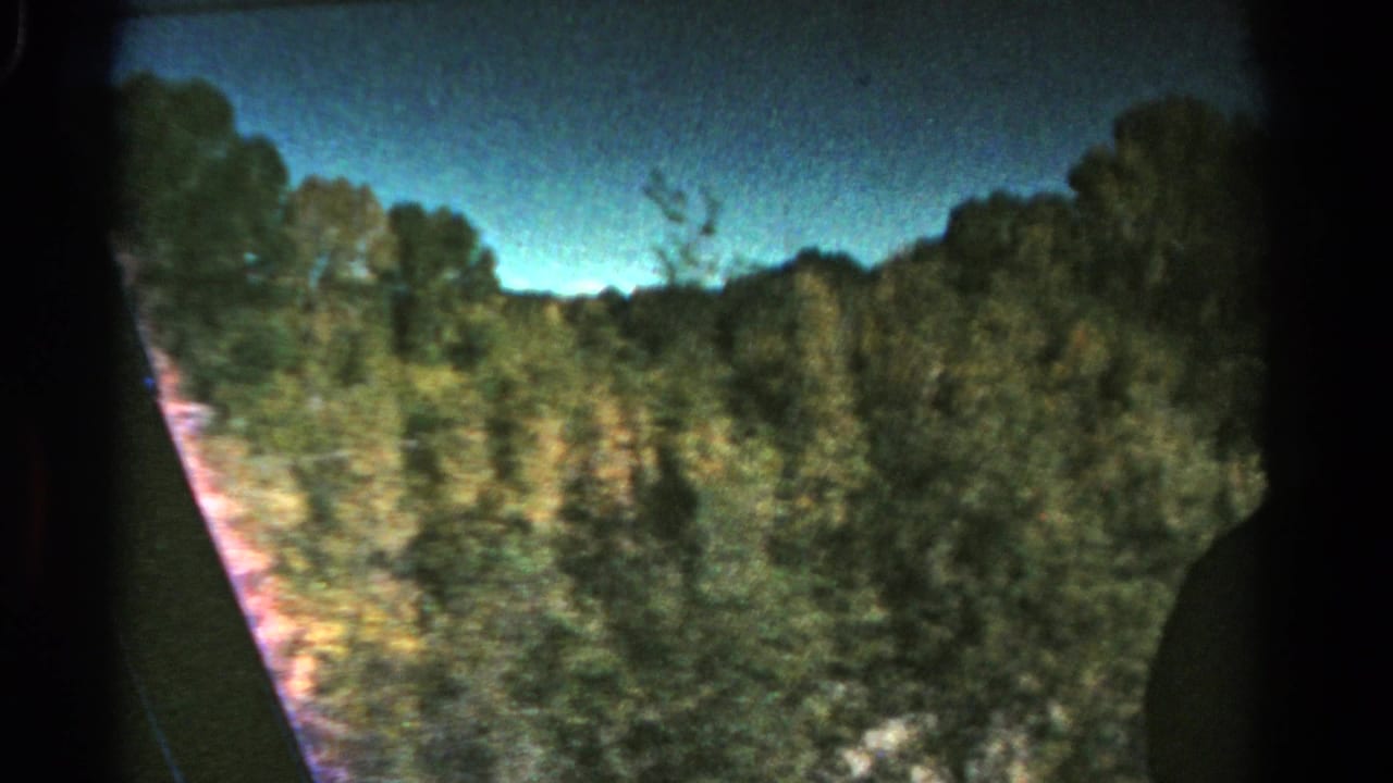1967 Colorado 8mm frame showing trees, low hills, blue sky, and dark vehicle-window edges from a moving passenger view.